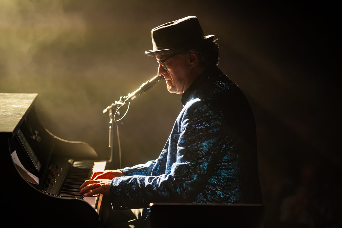Musiker am Klavier in schimmernder Jacke mit Mikrofon, umgeben von sanftem Licht.