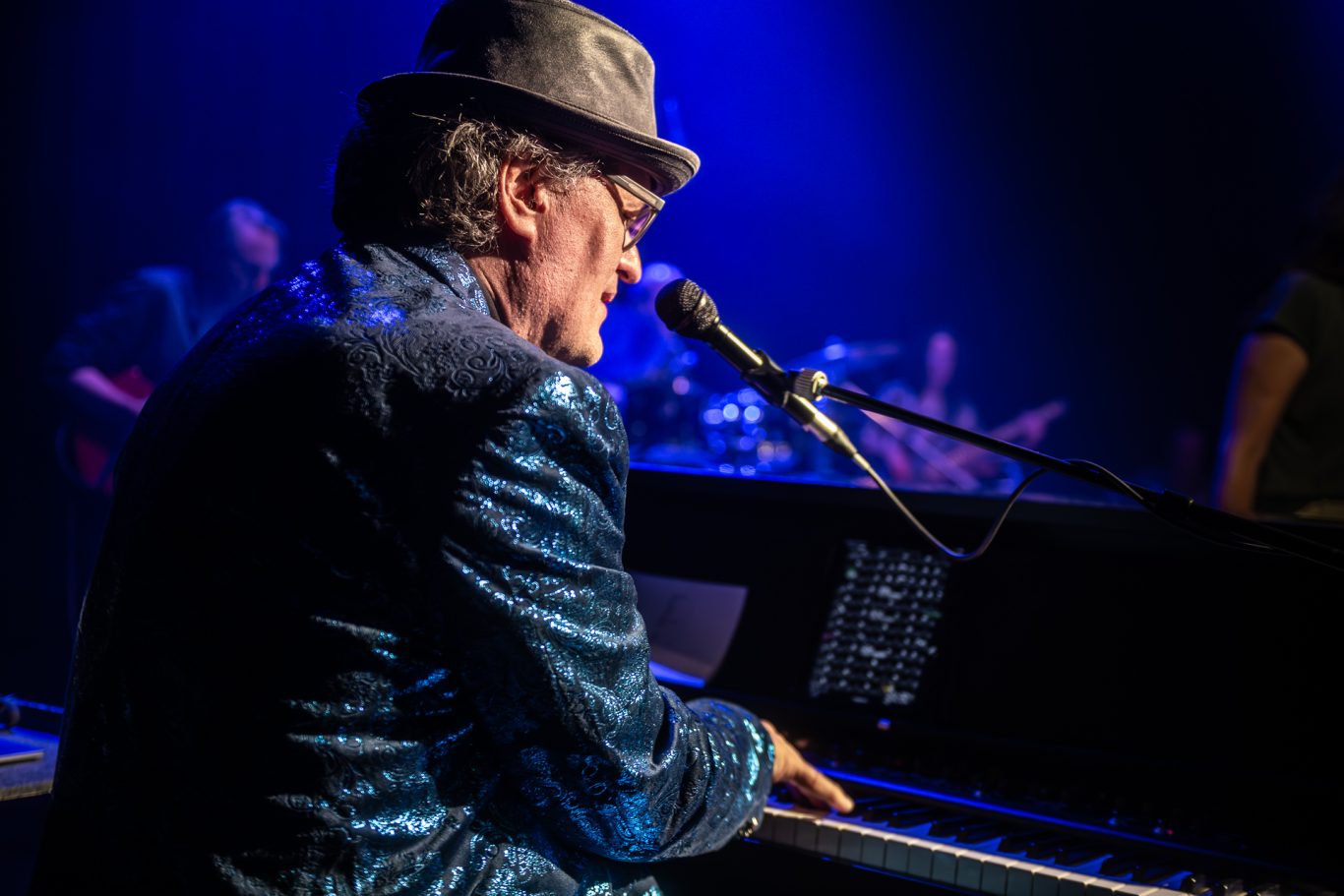 Mann mit Hut spielt Piano auf der Bühne, beleuchtet von blauer Lichtstimmung.