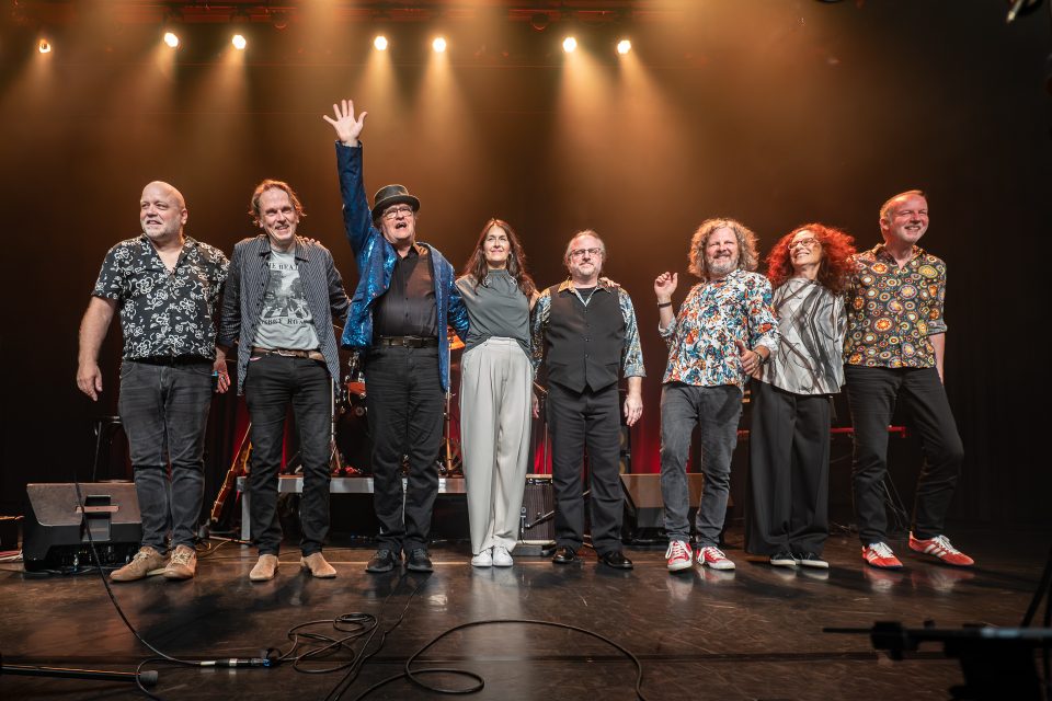 Eine Gruppe von acht Musikern steht auf der Bühne und begrüßt das Publikum.