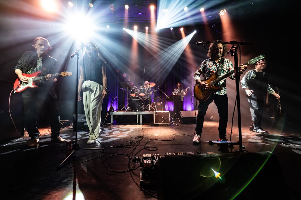 Band spielt live auf einer Bühne mit farbigen Lichtern und Instrumenten.