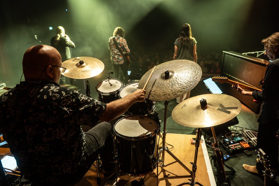 Ein Drummer spielt auf der Bühne, im Hintergrund stehen Musiker und eine Menschenmenge.