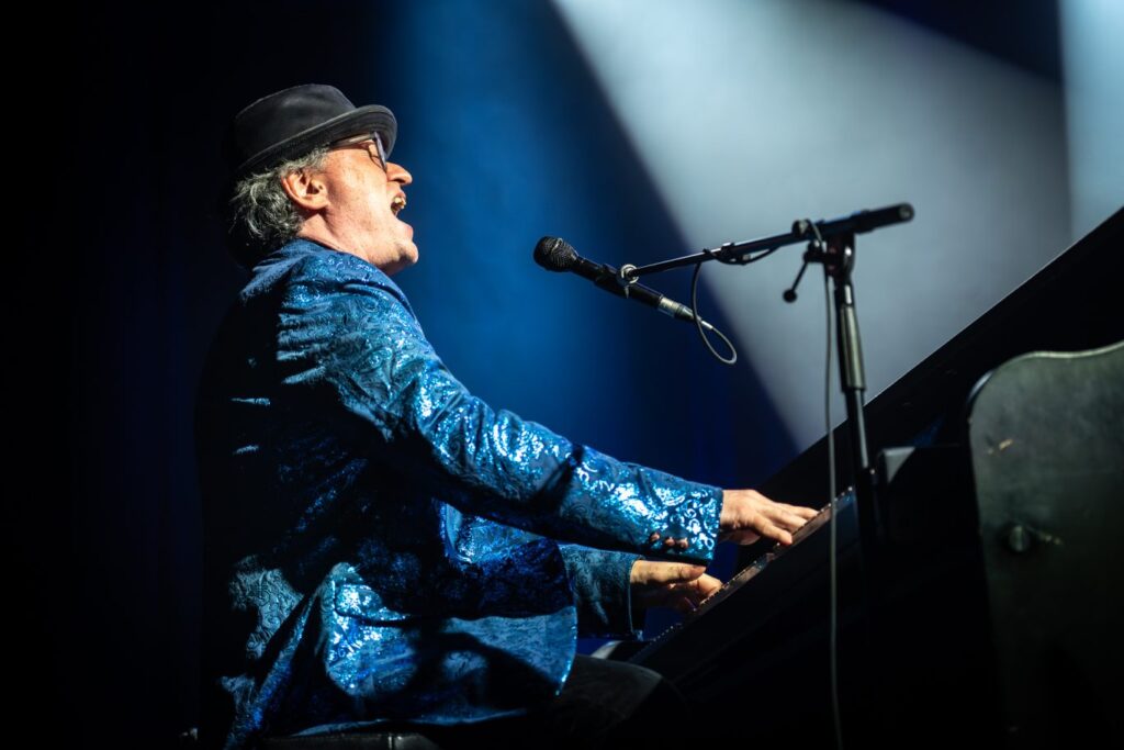 Ein Musiker in glitzerndem Anzug spielt begeistert Klavier und singt.