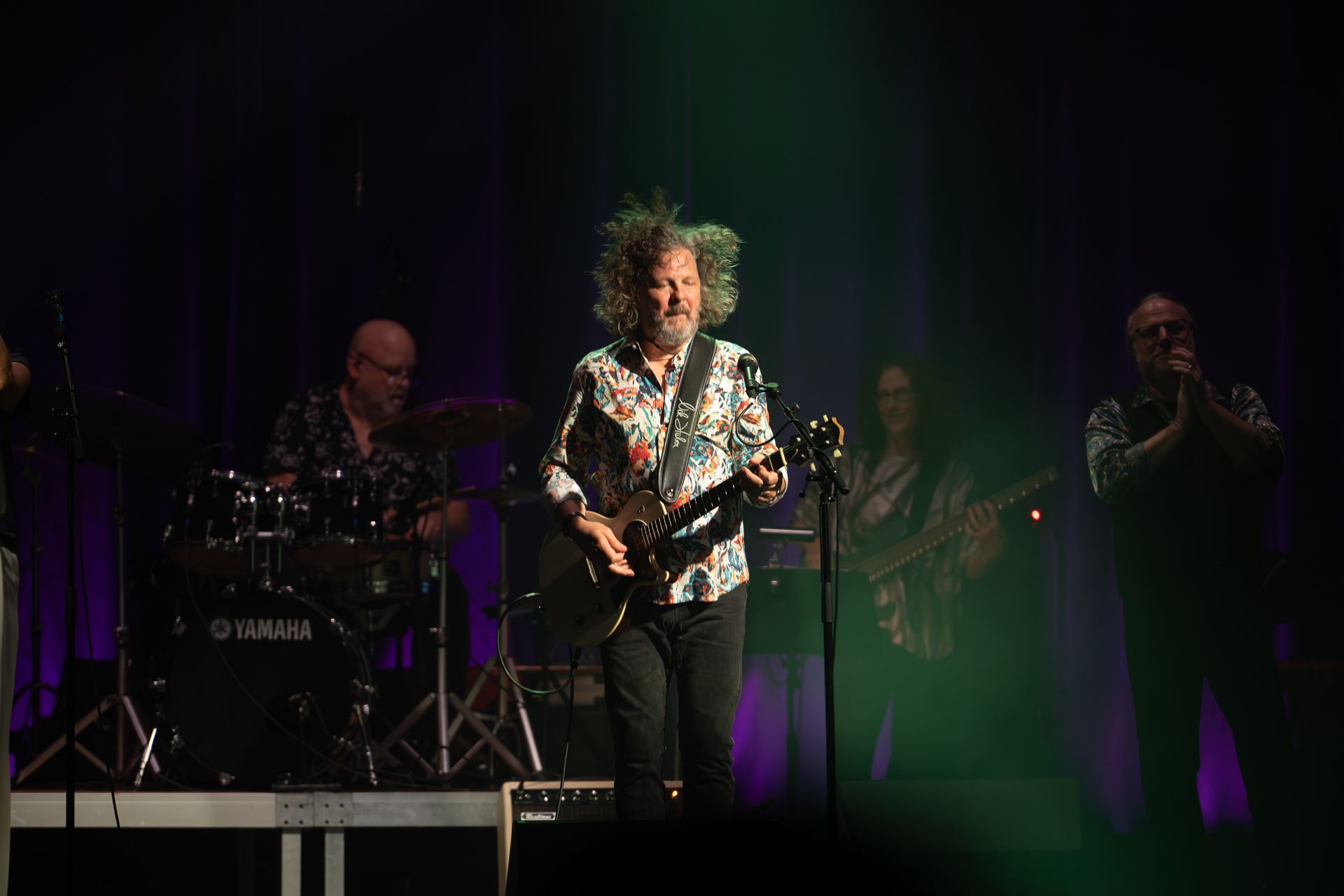 Musiker mit Gitarre auf der Bühne, umgeben von Bandmitgliedern im Hintergrund.