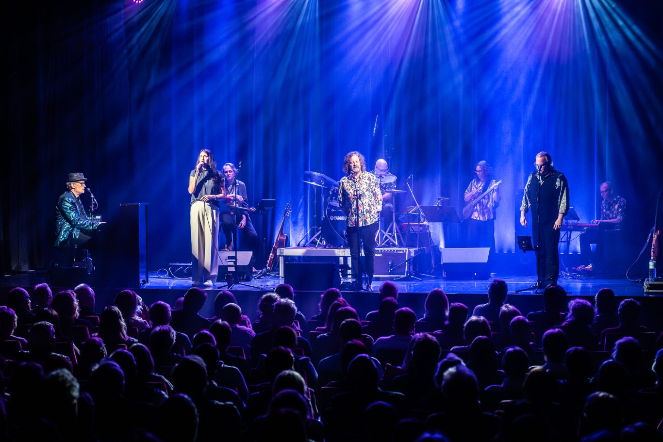 Live-Musikaufführung auf der Bühne mit einer Band und einem Publikum im Vordergrund.